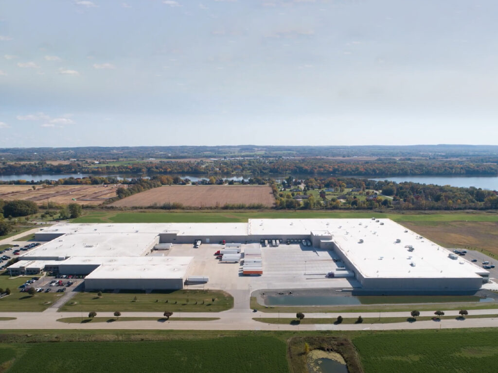 Aerial view of Green Bay Packaging’s Folding Carton Division facility, featuring large white-roofed buildings, loading docks, and surrounding green fields near a river.