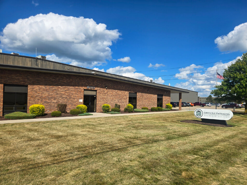 Exterior view of Green Bay Packaging's Archbold Division facility with brick building, company sign, and landscaped lawn under a blue sky with clouds.