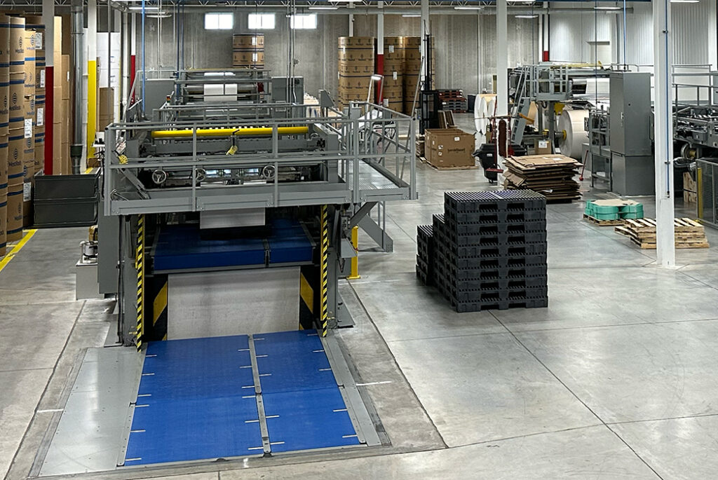 Sheeter machine inside Green Bay Packaging facility, featuring industrial rollers, metal framework, blue conveyor platform, and stacked black pallets on a polished concrete floor.