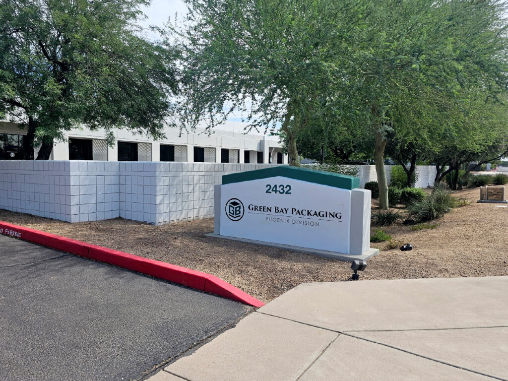 Outdoor view of the entrance to Green Bay Packaging’s Phoenix Division. The image shows a white and green sign with the company logo and address number “2432” in front of a low white block wall. Behind the sign are several green trees and shrubs, with a white building visible in the background. The ground around the sign and plants is covered with gravel, and a red-painted curb lines the edge of the asphalt road. The sky is clear and sunny